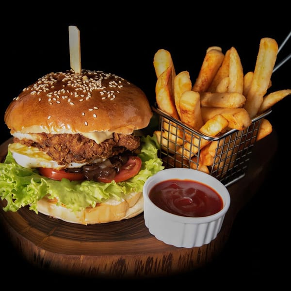 Crispy chicken burger with fried egg, lettuce, tomato, and fries with ketchup on a wooden board.