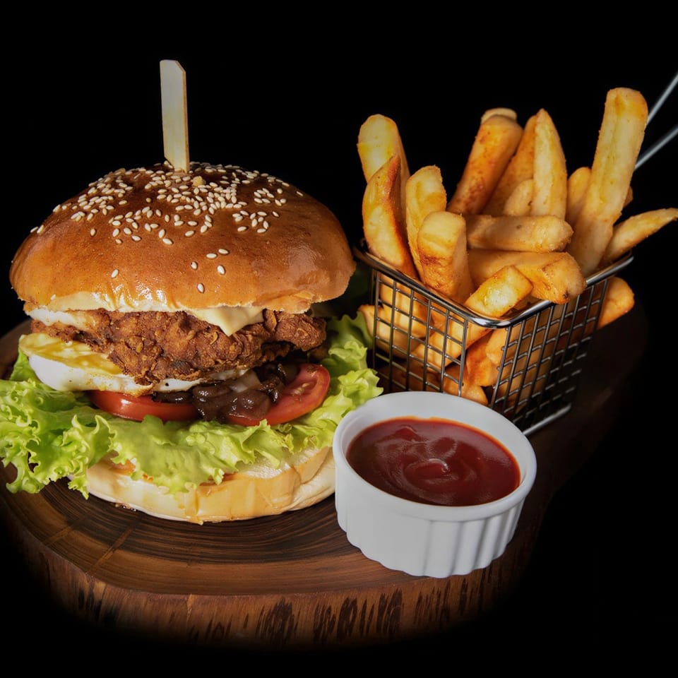 Crispy chicken burger with fried egg, lettuce, tomato, and fries with ketchup on a wooden board.